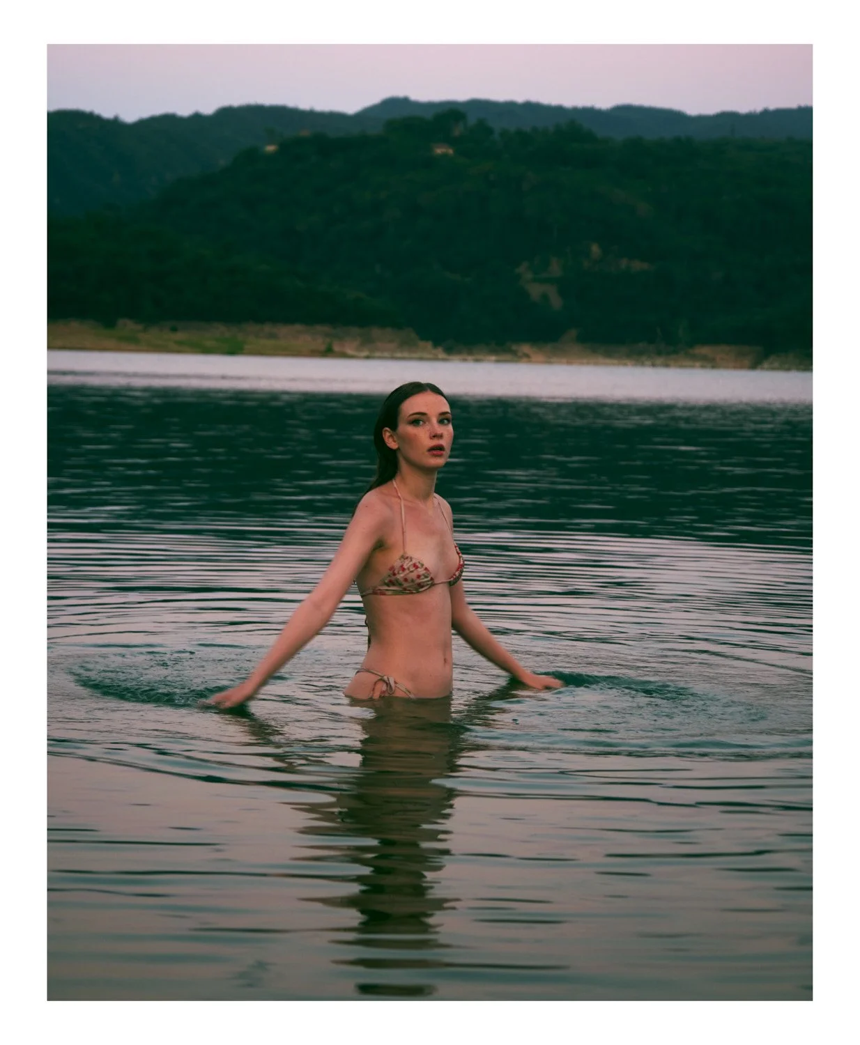 A woman in a bikini standing in a calm body of water with mountains in the background during dusk or dawn.