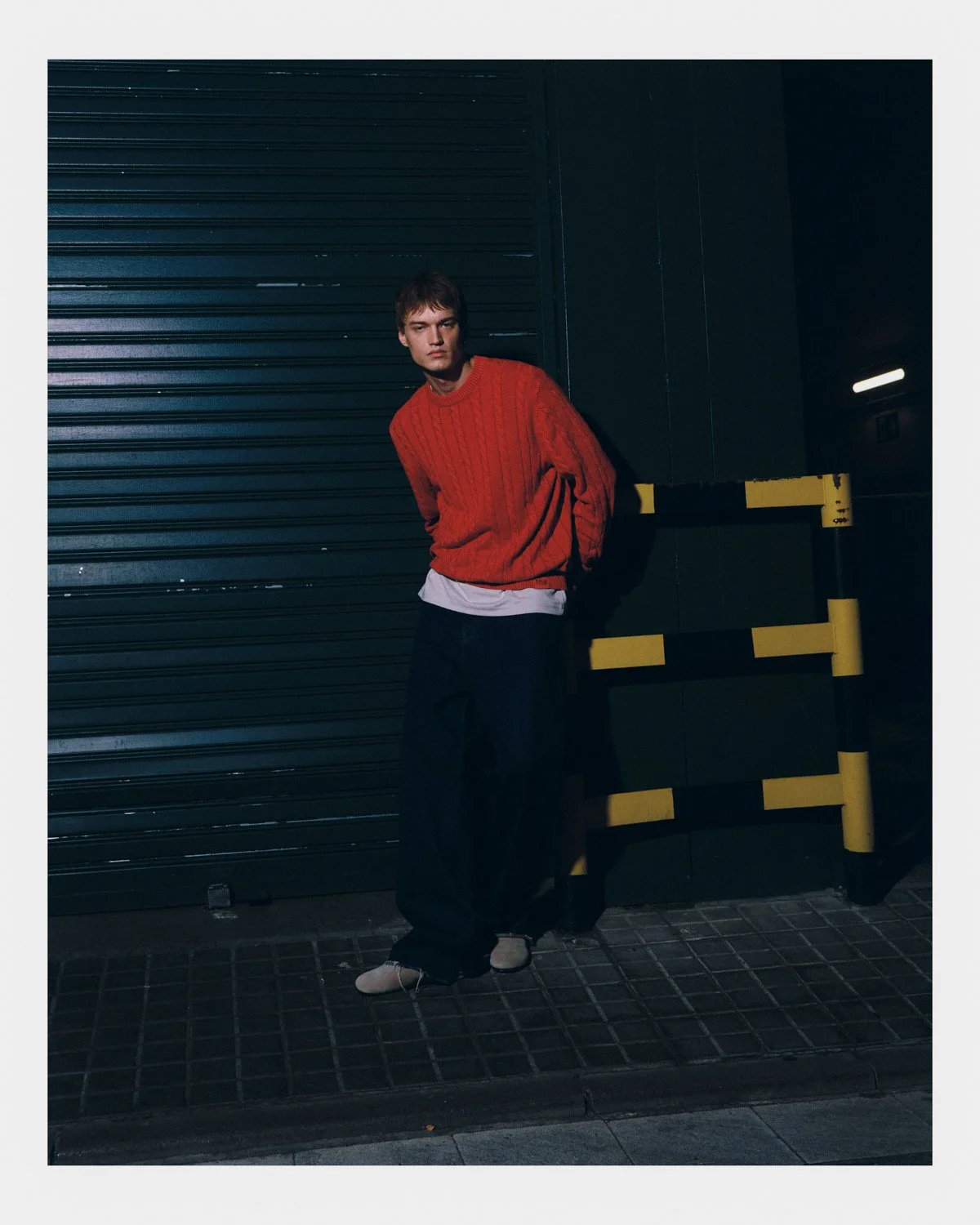 A young man in a red sweater and black pants leaning against a dark wall in an urban setting during nighttime.