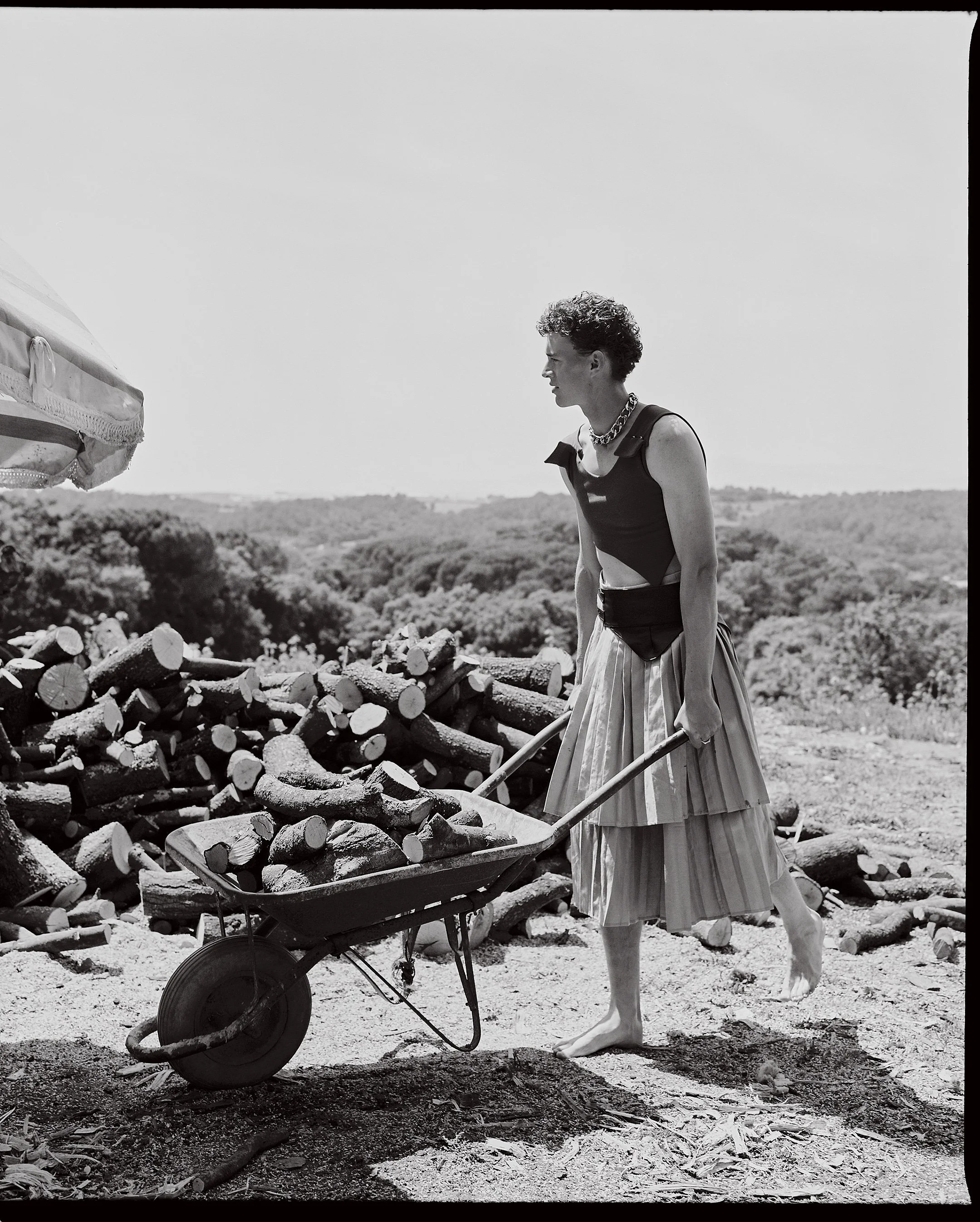 A woman with curly hair, dressed in a sleeveless top and long pleated skirt, pushes a wheelbarrow filled with logs outdoors, with a background of trees and an open sky.