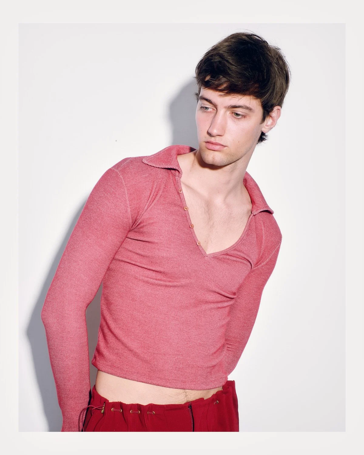 A young man with dark hair wearing a pink long-sleeve shirt with buttons and red pants, standing against a white background.