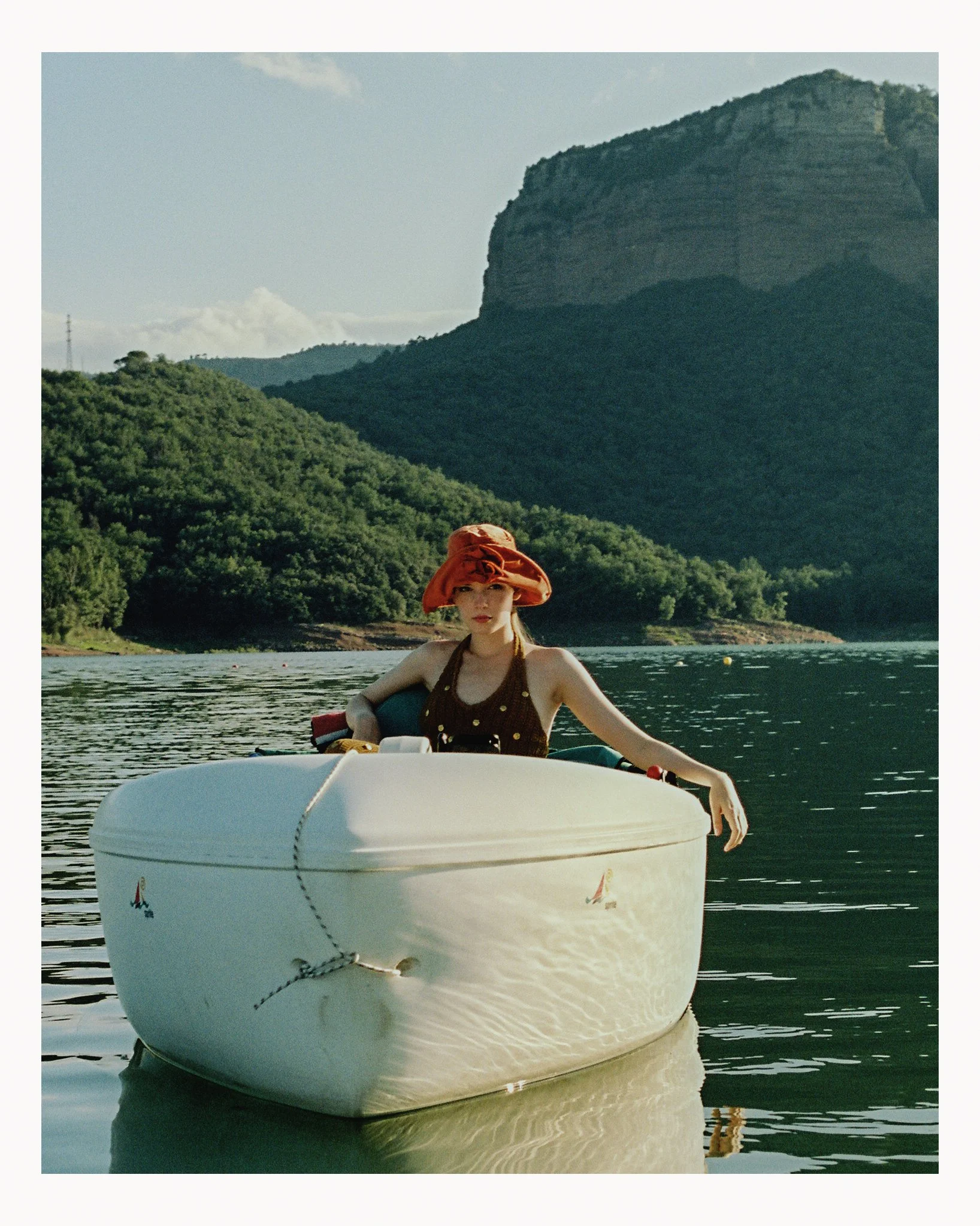 A woman in a brown dress and orange hat sits in a small white boat on a calm lake, with forested hills and a large rocky mountain in the background.