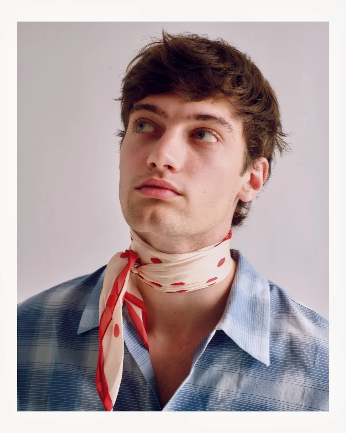 Young man with a thoughtful expression, wearing a blue plaid shirt and a red polka dot scarf around his neck.