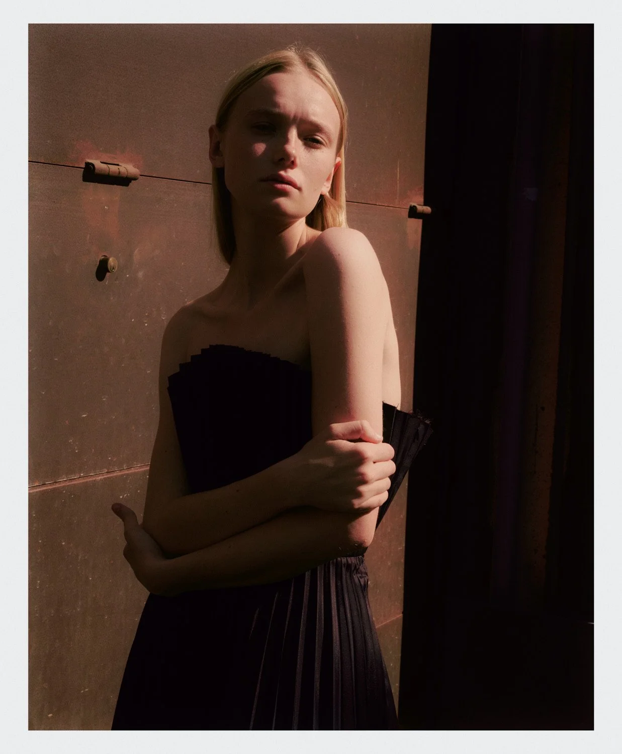 A young woman with blonde hair, wearing a strapless black dress, stands against a brown background with her arms crossed, looking at the camera with a serious expression.