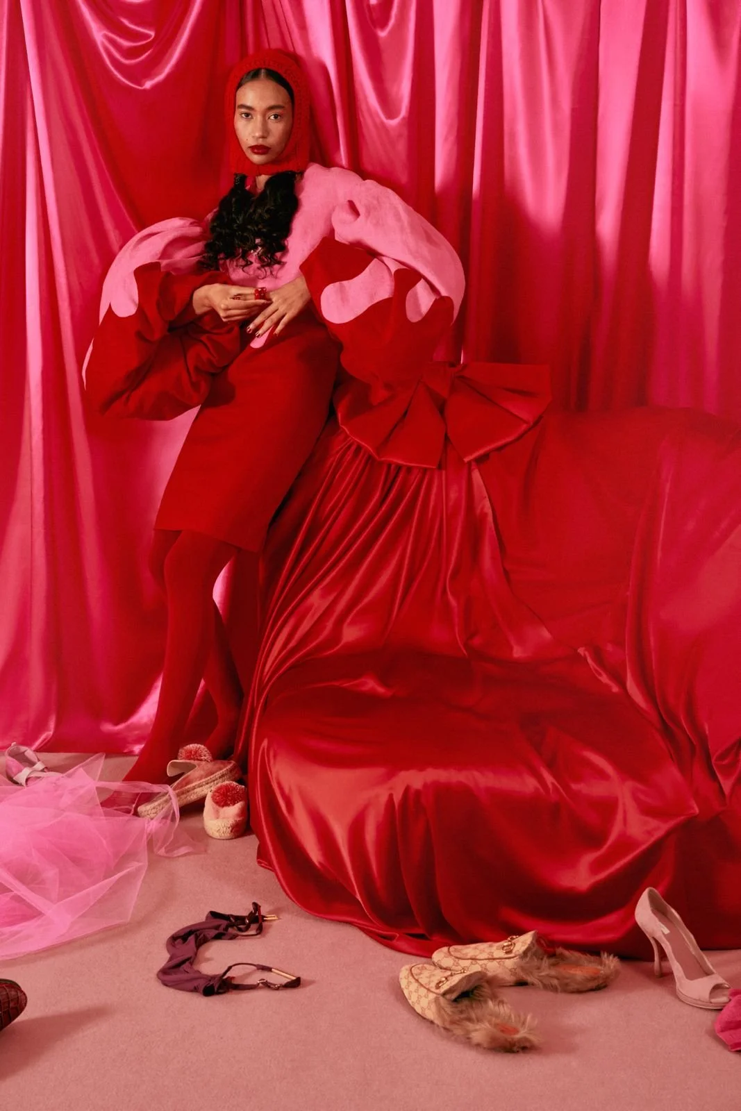 A woman dressed in a pink and red outfit standing in front of pink satin curtains with a large red satin satin drape and bow on the side, surrounded by various shoes and accessories on a pink carpet.