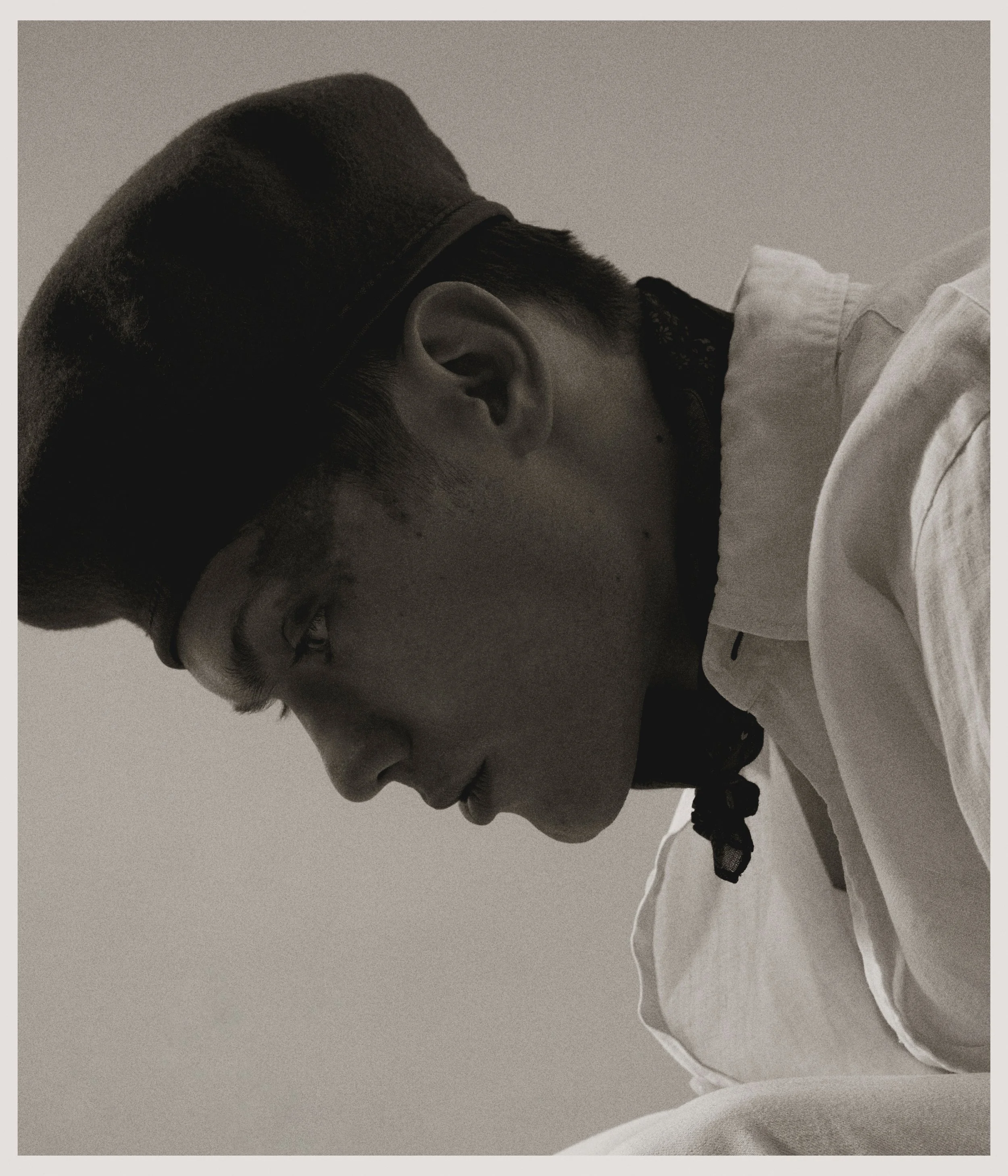 A black and white side profile of a young man wearing a cap, a collared shirt, and a neck scarf, looking downward with a serious expression.
