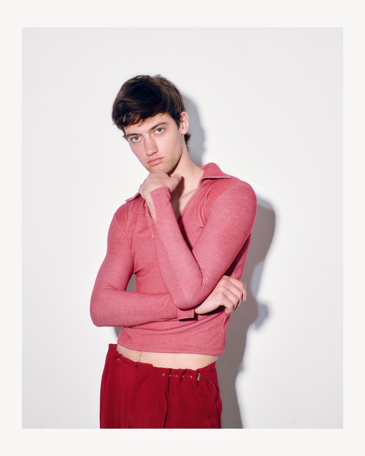 Young man with dark hair wearing a pink long-sleeve top and red pants standing against a white background.