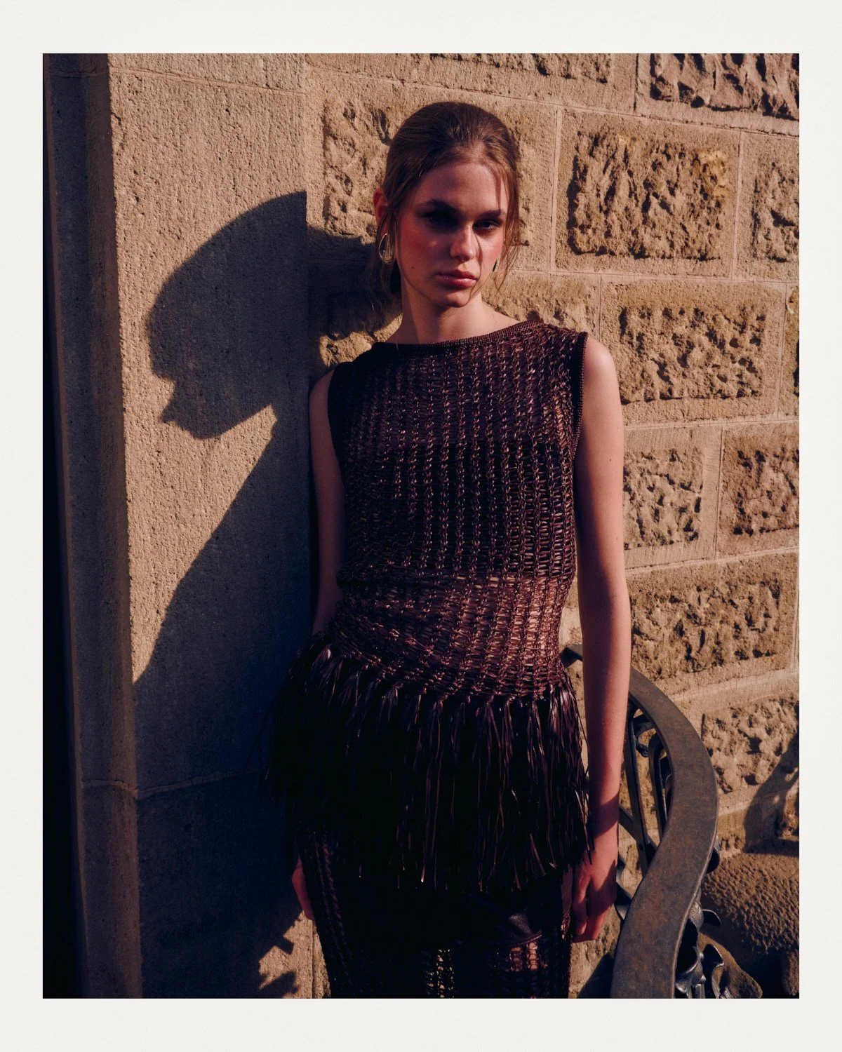 A woman with brown hair in a bun wearing a dark, textured sleeveless dress, standing outdoors against a stone wall, with a shadow cast on the wall behind her.