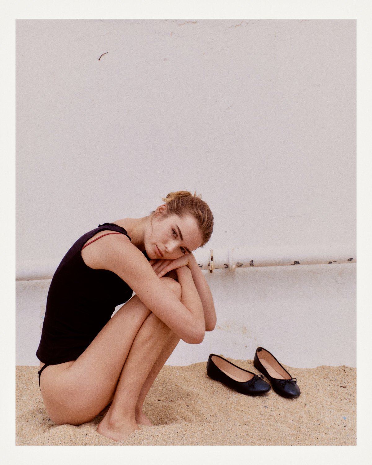 A woman with red hair in a black dress sits on sand against a white wall, hugging her knees and looking at the camera, with black ballet flats on the sand nearby.