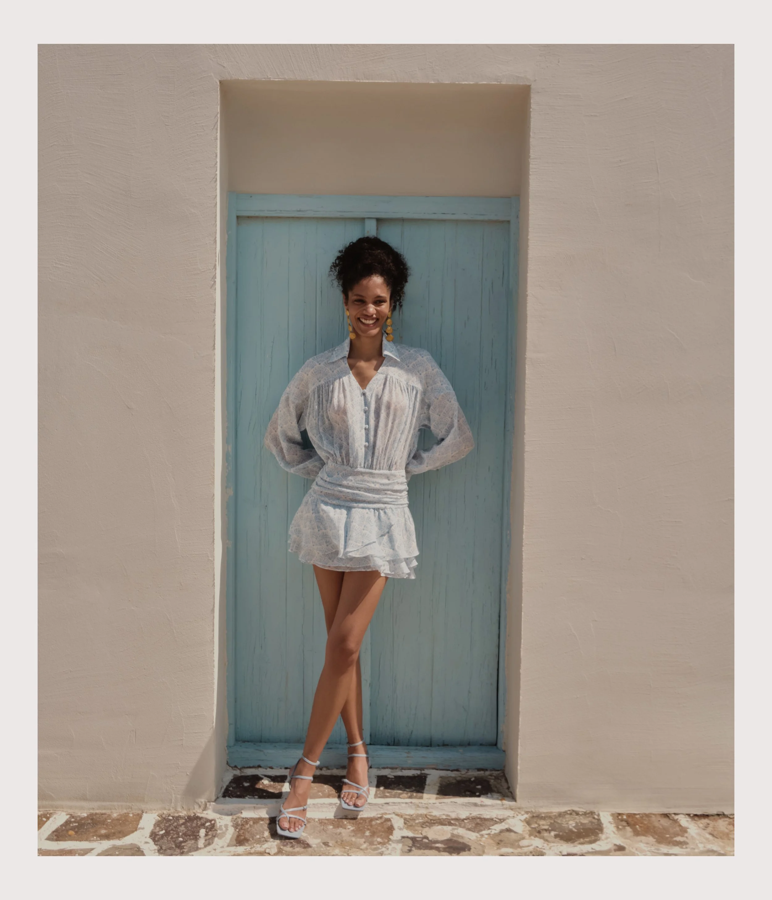 A woman standing in front of a light blue door, smiling, wearing a white dress with sheer long sleeves and white high-heeled sandals.