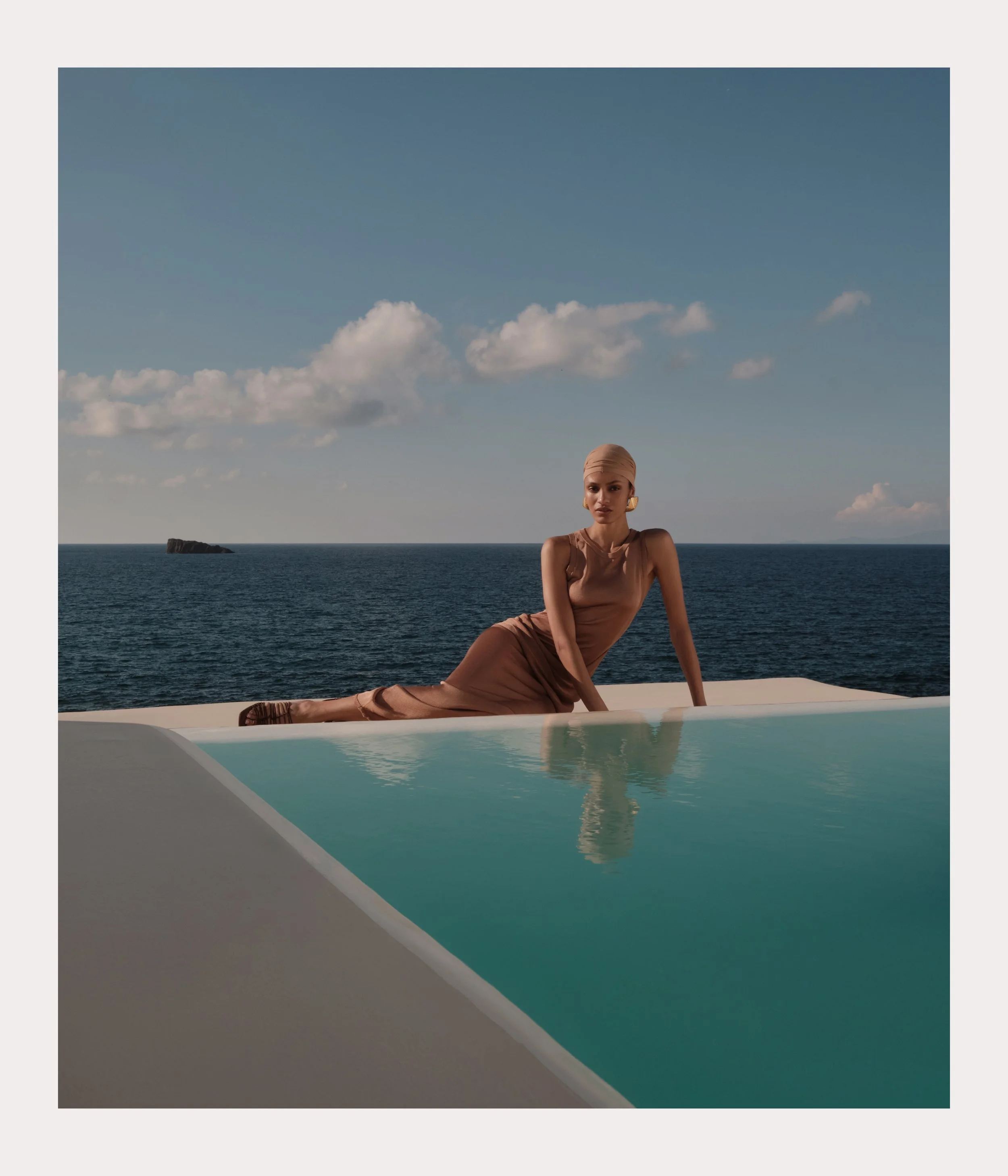 A woman in a tan dress posing by a pool with the ocean and a cloudy sky in the background.