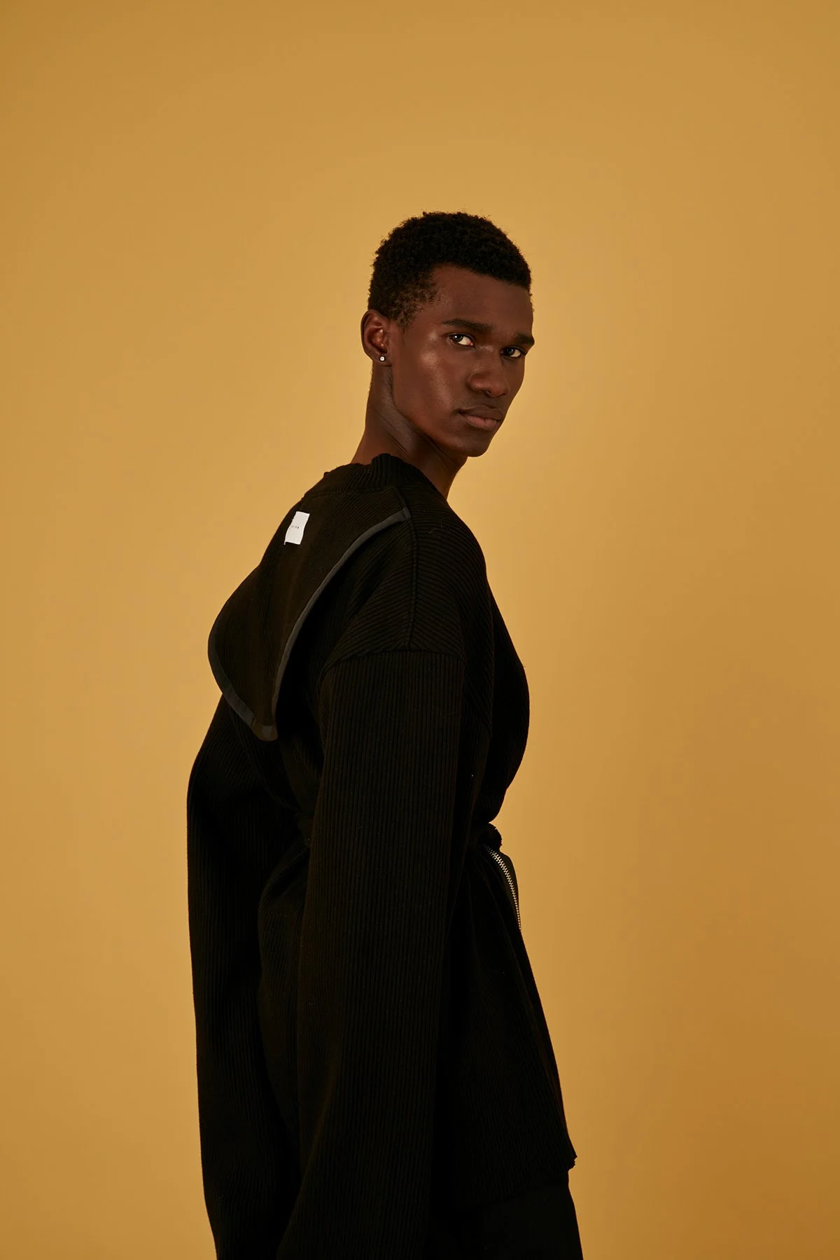 A young man with dark skin and short curly hair stands against a yellow background, looking over his shoulder at the camera, wearing a black zip-up jacket.