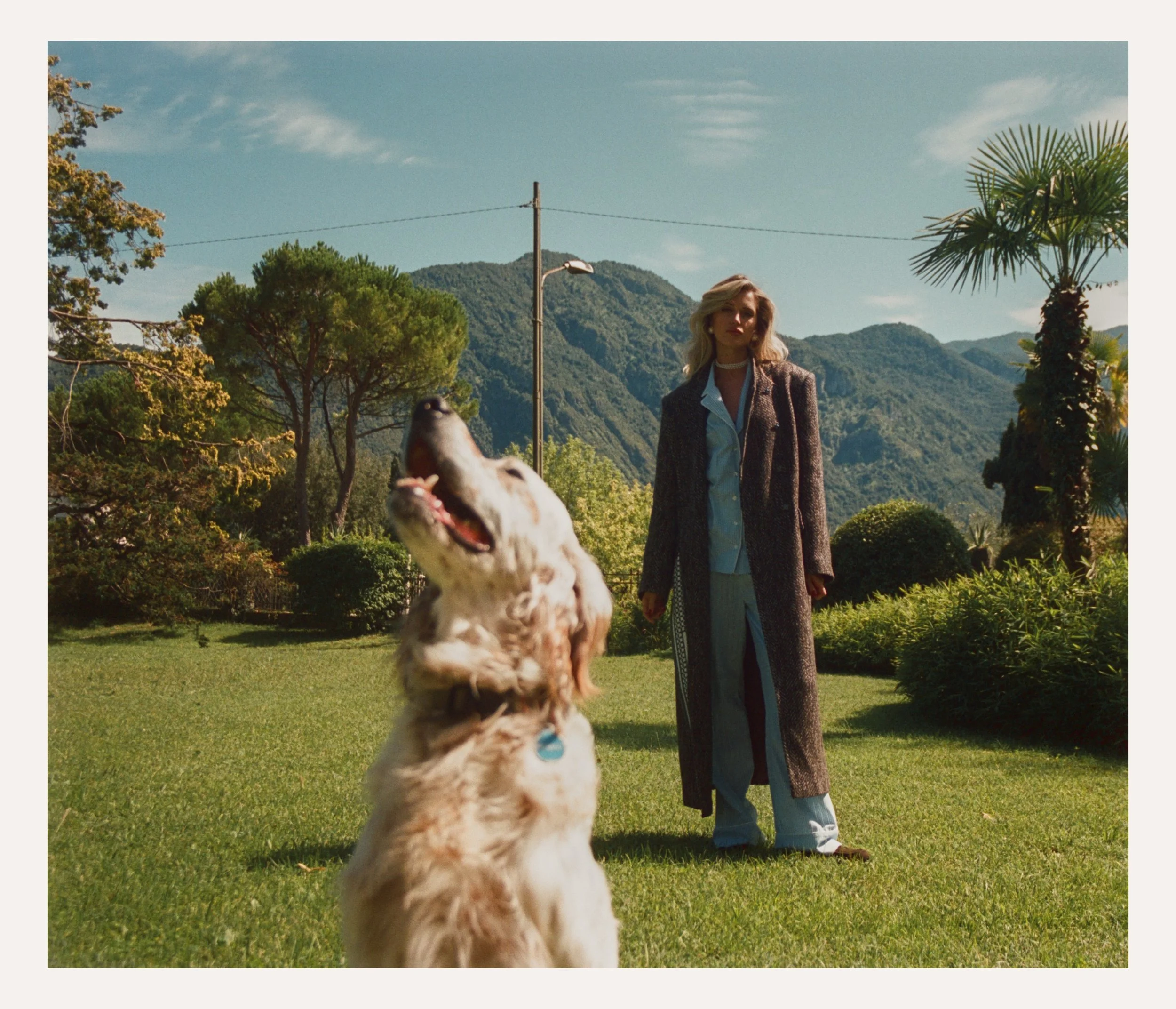 A woman with blonde hair in a long brown coat standing on a green lawn with a happy dog in the foreground and mountains in the background under a partly cloudy sky.