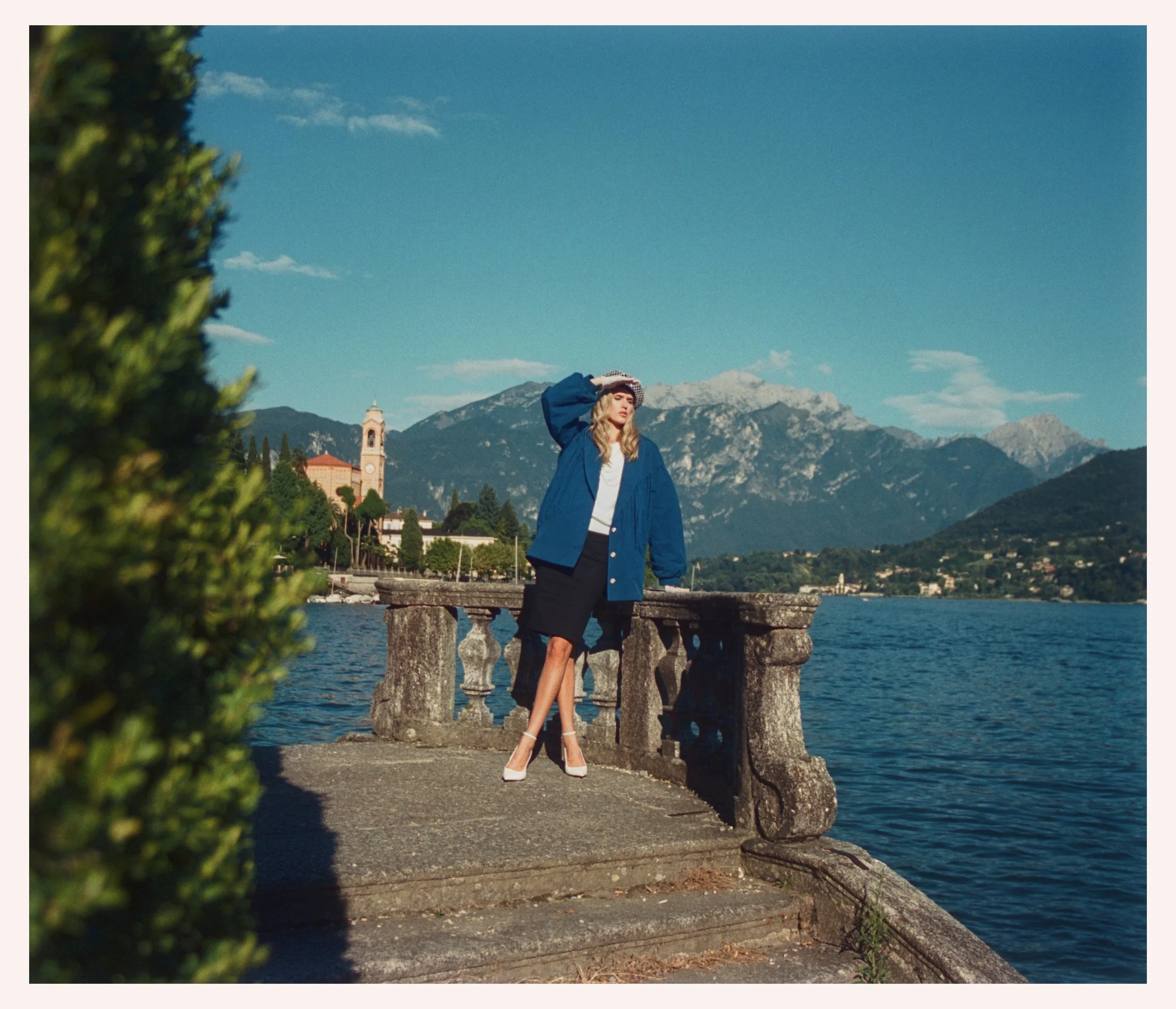 A woman in a blue coat, white heels, and a hat standing on a stone promenade near a body of water with mountains in the background.