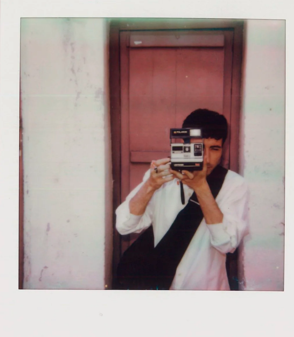 Young man taking a mirror selfie with a vintage instant camera in front of a pink door.