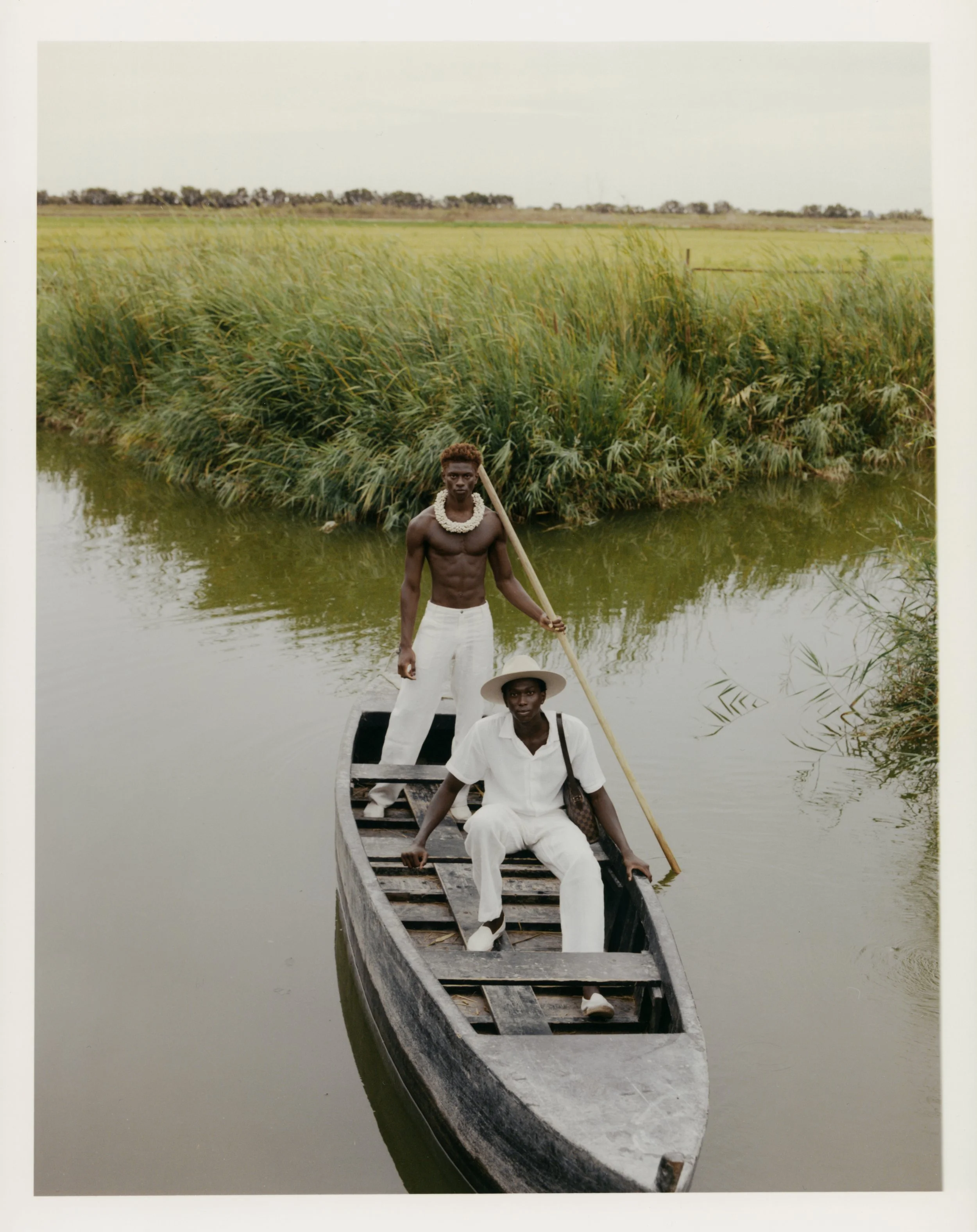 Two men on a small wooden boat in a river, with tall grass and open field in the background.