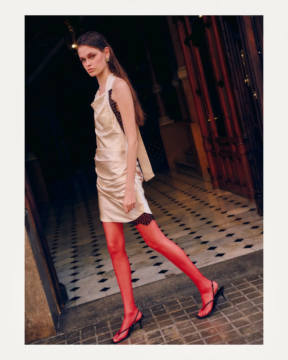 A young woman in a shiny cream dress, red stockings with lace tops, and black high heels, standing outside a building at night.