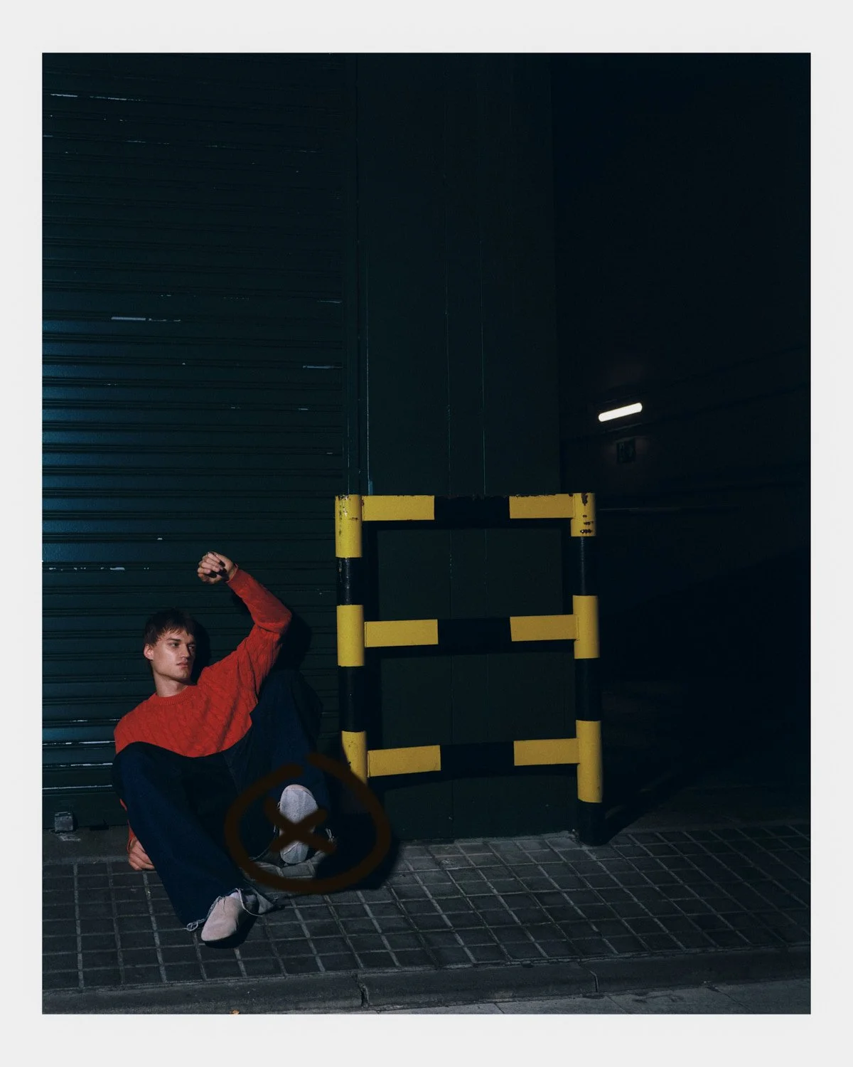 A young man sitting on the ground next to a dark, industrial building with a yellow and black barrier, wearing a red sweater and black pants, looking to the side in an urban setting at night.