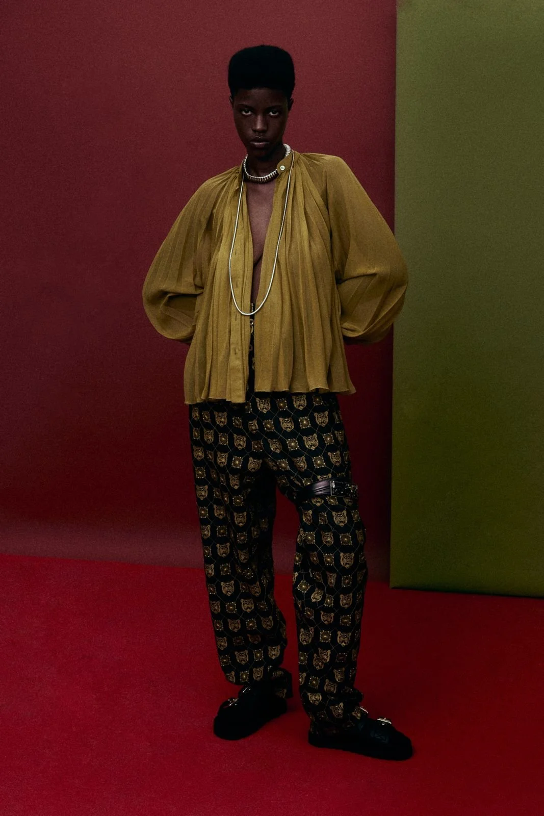 Fashion model wearing a mustard yellow pleated blouse, black patterned pants with gold designs, black shoes, layered necklaces, and a bracelet, standing against a multi-colored background with red and green sections.