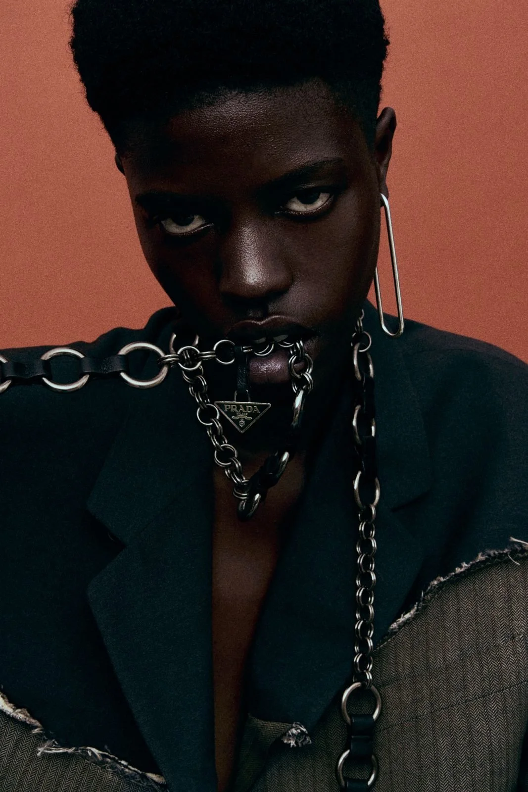 A Black person with short hair wears dark clothing and multiple silver chains around their neck and shoulders, with a Prada logo pendant, against a solid reddish-brown background.