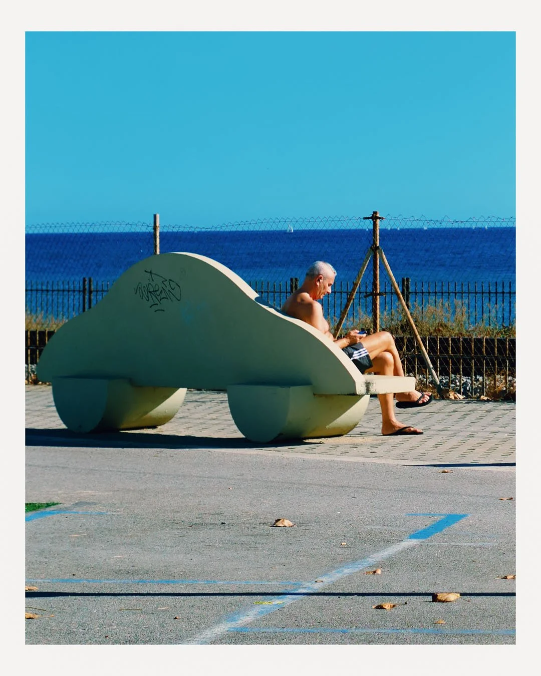 A man with gray hair sitting on a modern concrete bench shaped like a whale, near a fence and overlooking the ocean in bright, sunny weather.