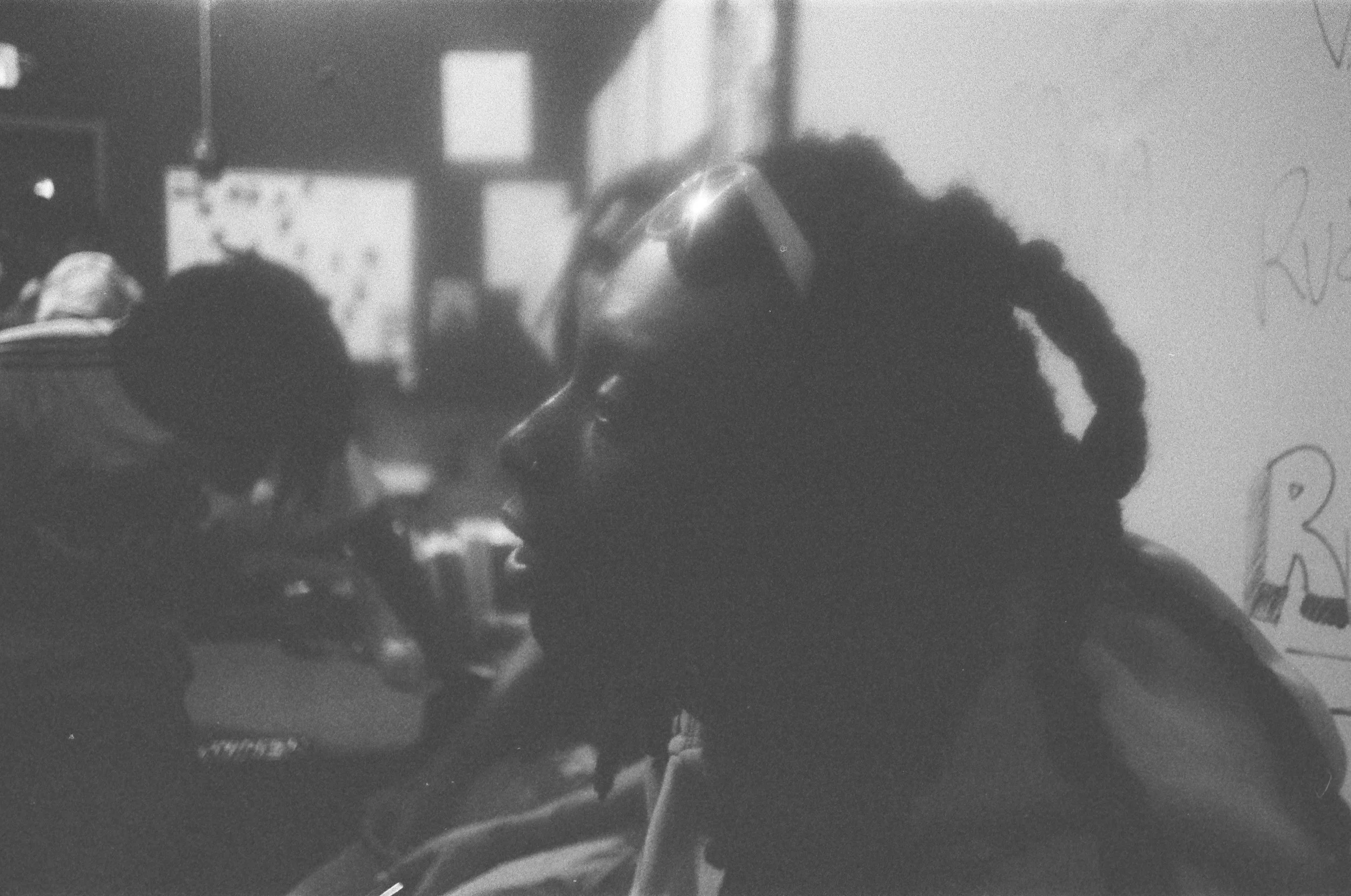 Profile of a person with dreadlocks wearing glasses, facing another individual with a cap, in a crowded indoor setting.