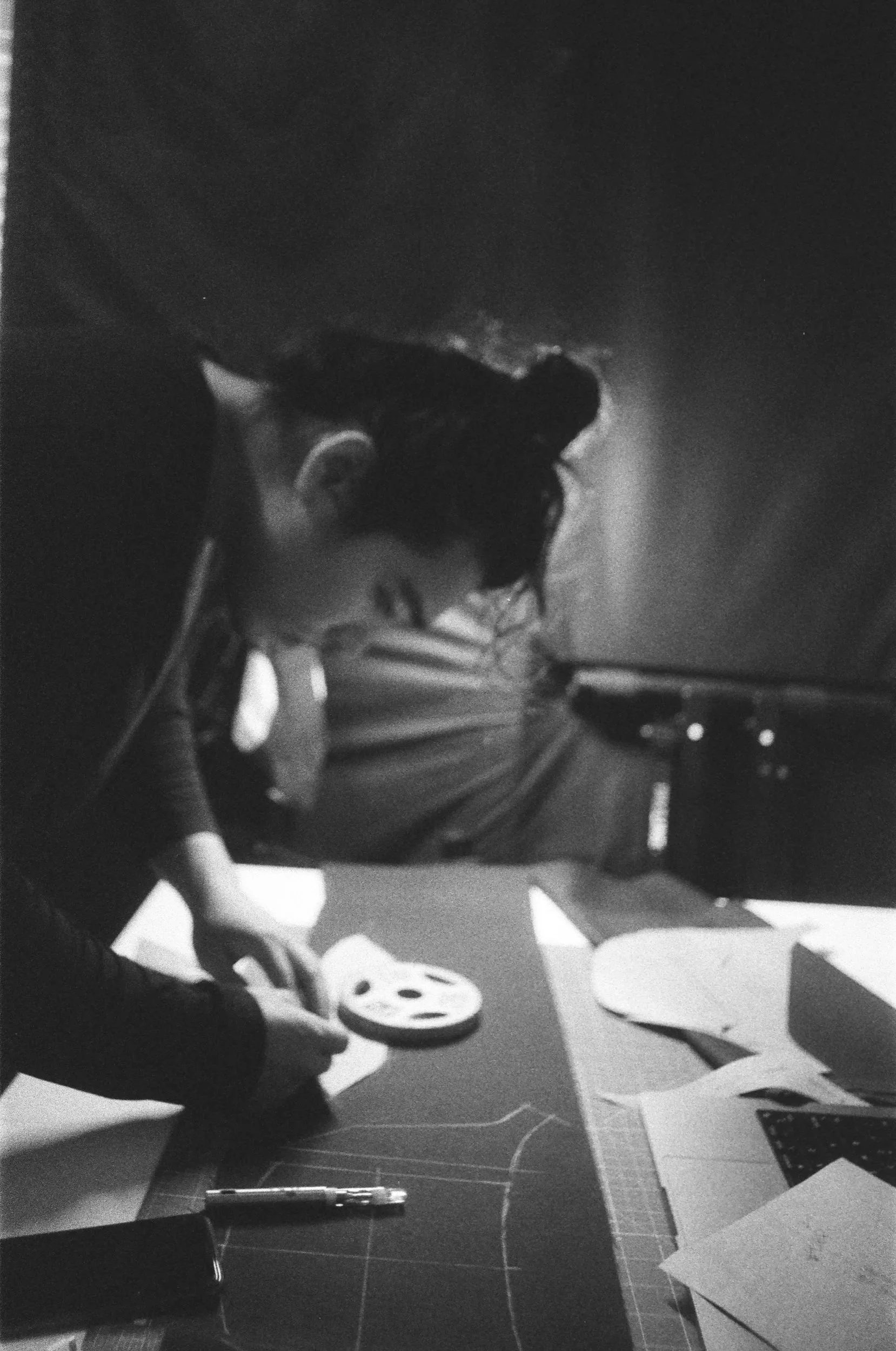 A person with long hair tied in a bun, working on a craft or project at a desk, with various papers and tools around.