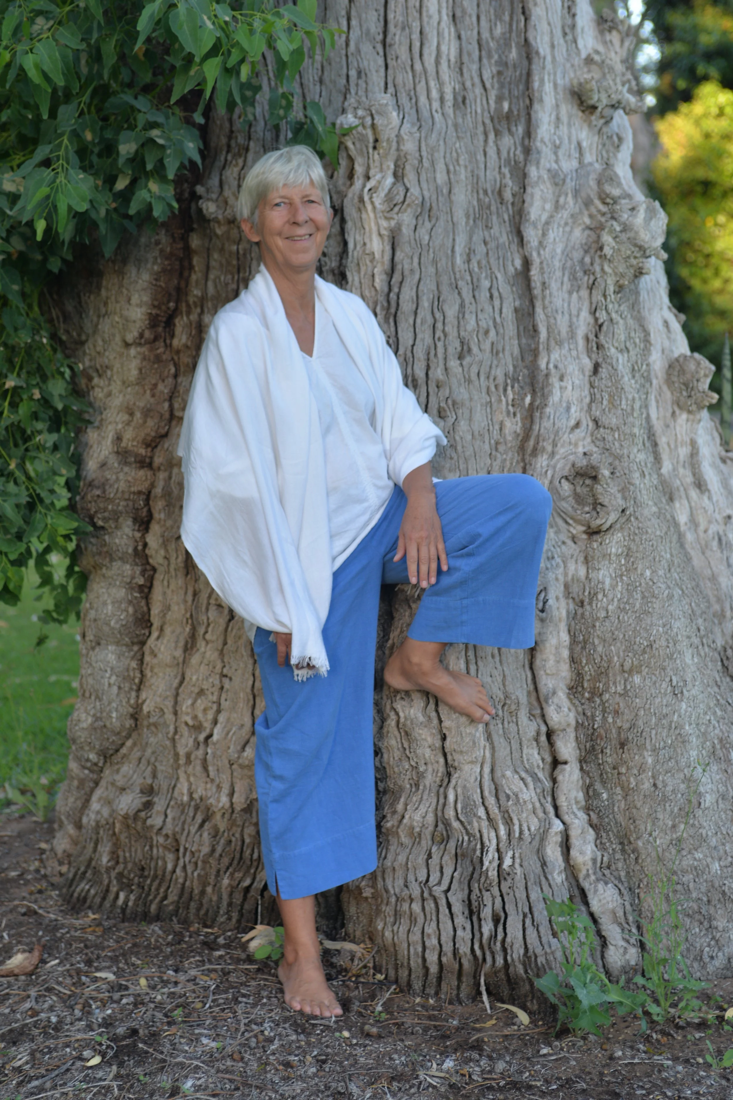 A smiling woman with short gray hair, dressed in white and blue, poses barefoot against a large tree trunk with green foliage around.
