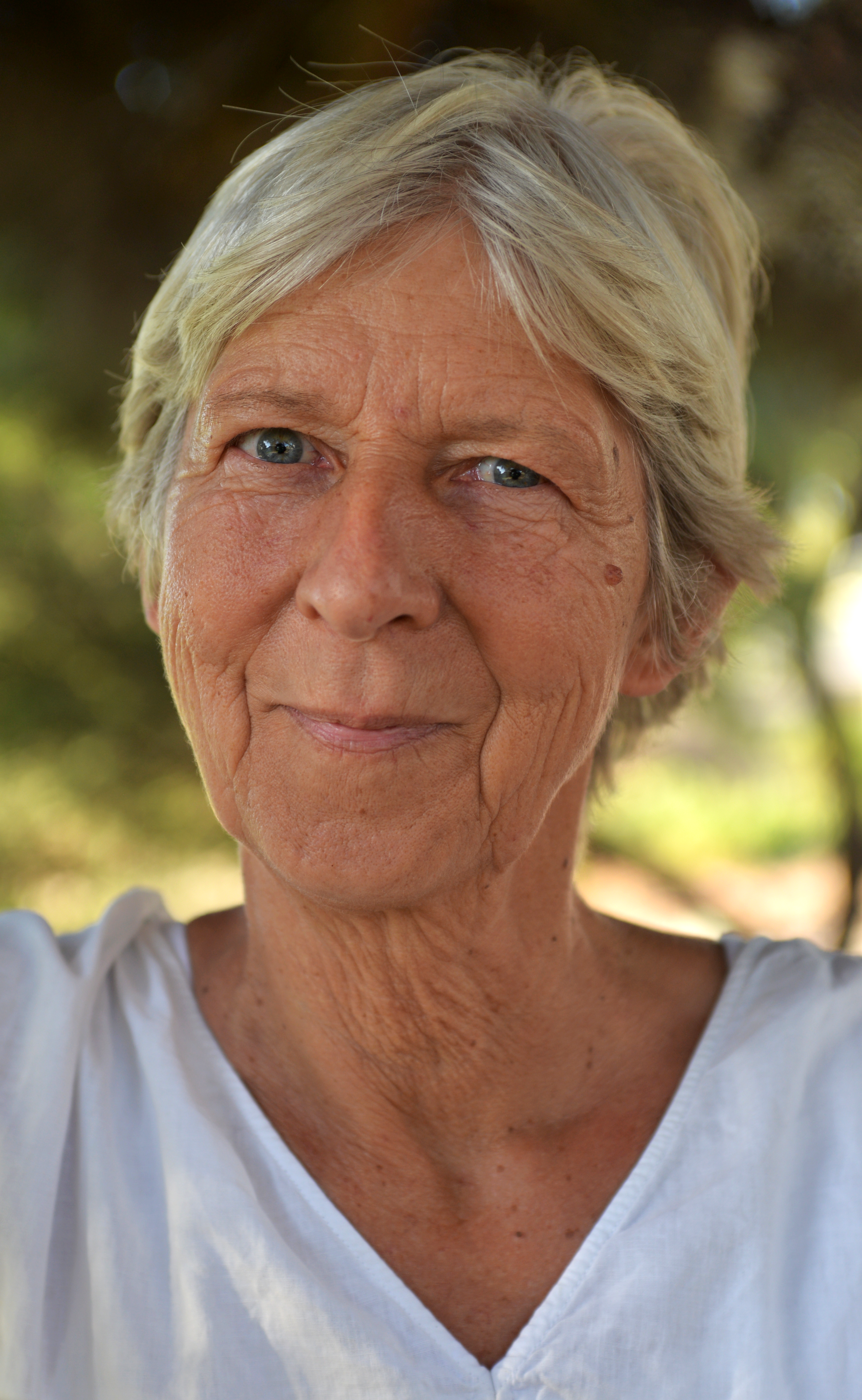 Close-up of an older woman with short, gray hair, blue eyes, and a gentle smile, standing outdoors with blurred trees and sunlight in the background.