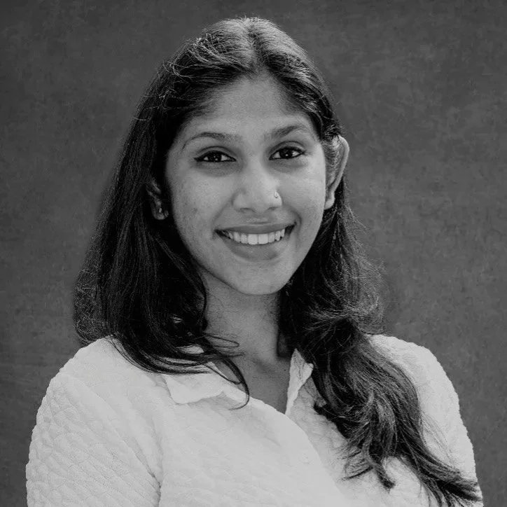 Black and white portrait of a young woman with long dark hair, smiling, wearing a light-colored textured top, standing against a plain dark background.