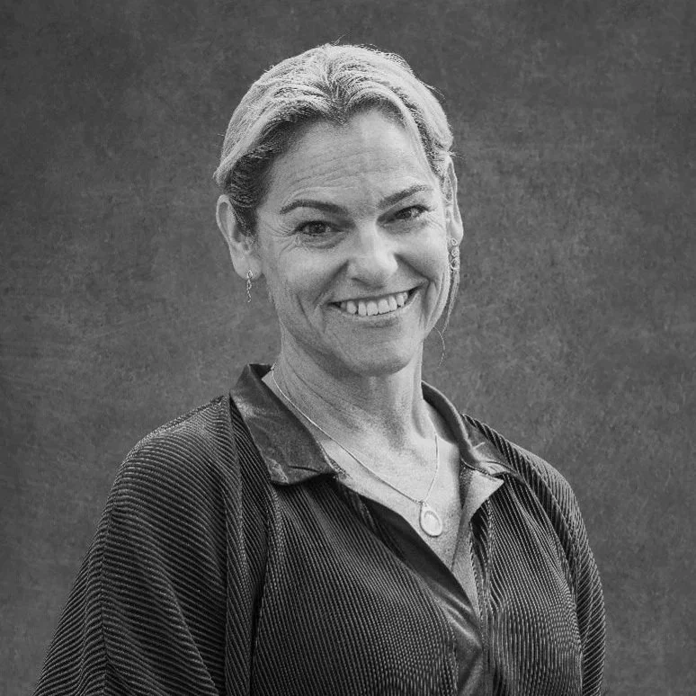 A smiling woman with short, light-colored hair, wearing a dark top and a necklace, standing against a textured background.