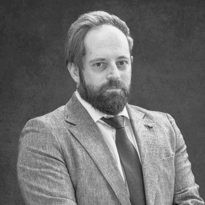 A man with a beard and mustache wearing a suit and tie, with arms crossed, looking at the camera against a dark background.