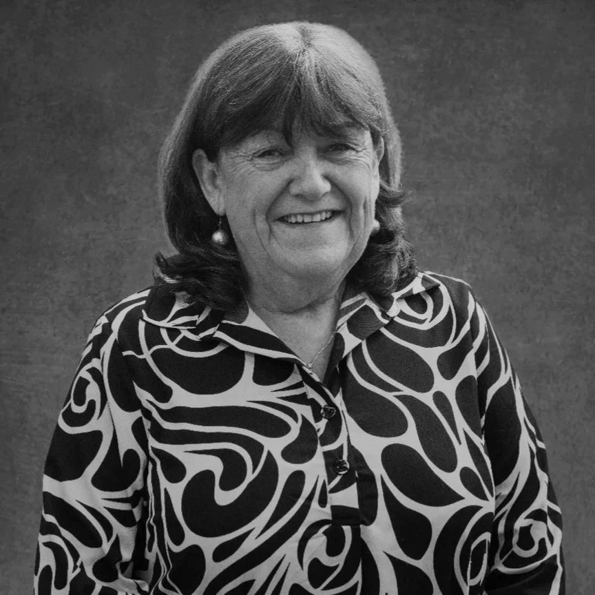 A smiling middle-aged woman with shoulder-length hair, wearing a patterned blouse with bold black and white swirling designs and pearl earrings, standing against a plain background.