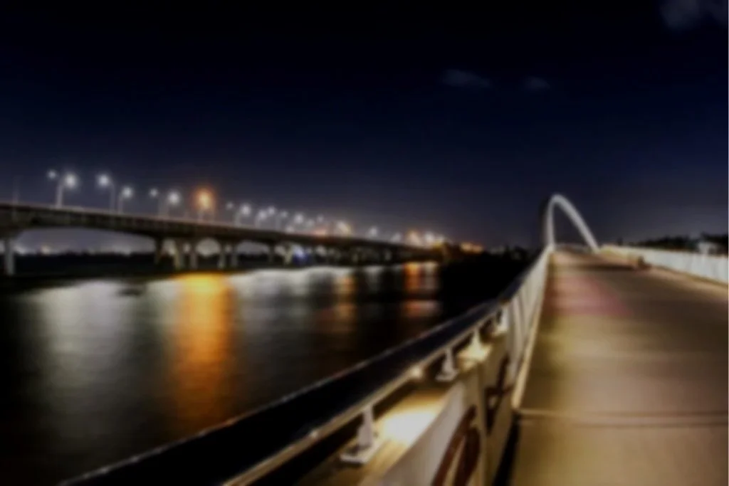 Night view of a riverwalk promenade with a modern arch bridge in the background, illuminated streetlights, and calm water reflecting city lights.