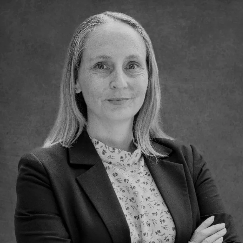 A woman with shoulder-length hair, wearing a blazer over a floral blouse, smiling with her arms crossed against a plain background.
