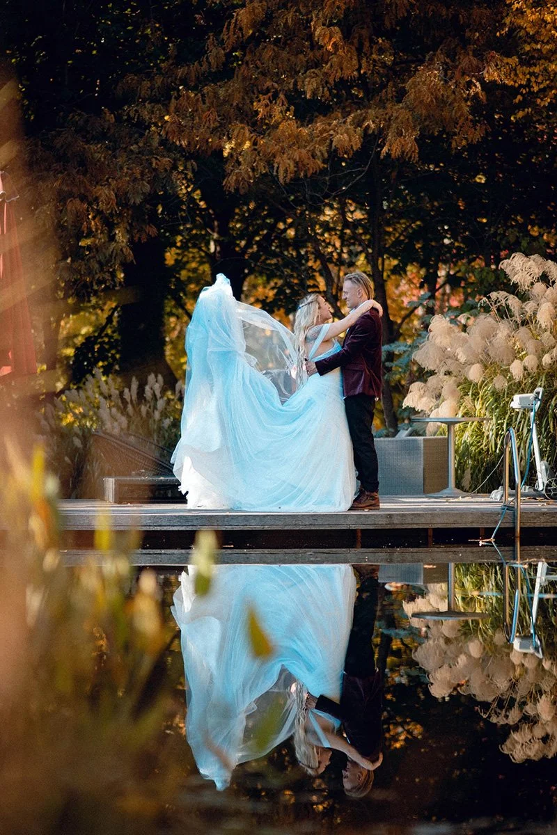 A bride and groom dancing on a wooden platform beside a natural pool at sunset, with the reflection of the couple visible in the water, surrounded by trees and tall grasses.