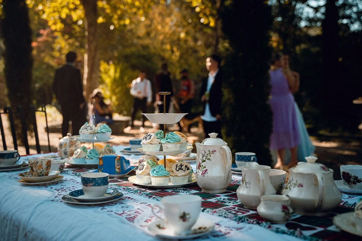 An outdoor tea party with a table set with teacups, teapots, and cupcakes, with people in formal attire in the background under trees with fall foliage.