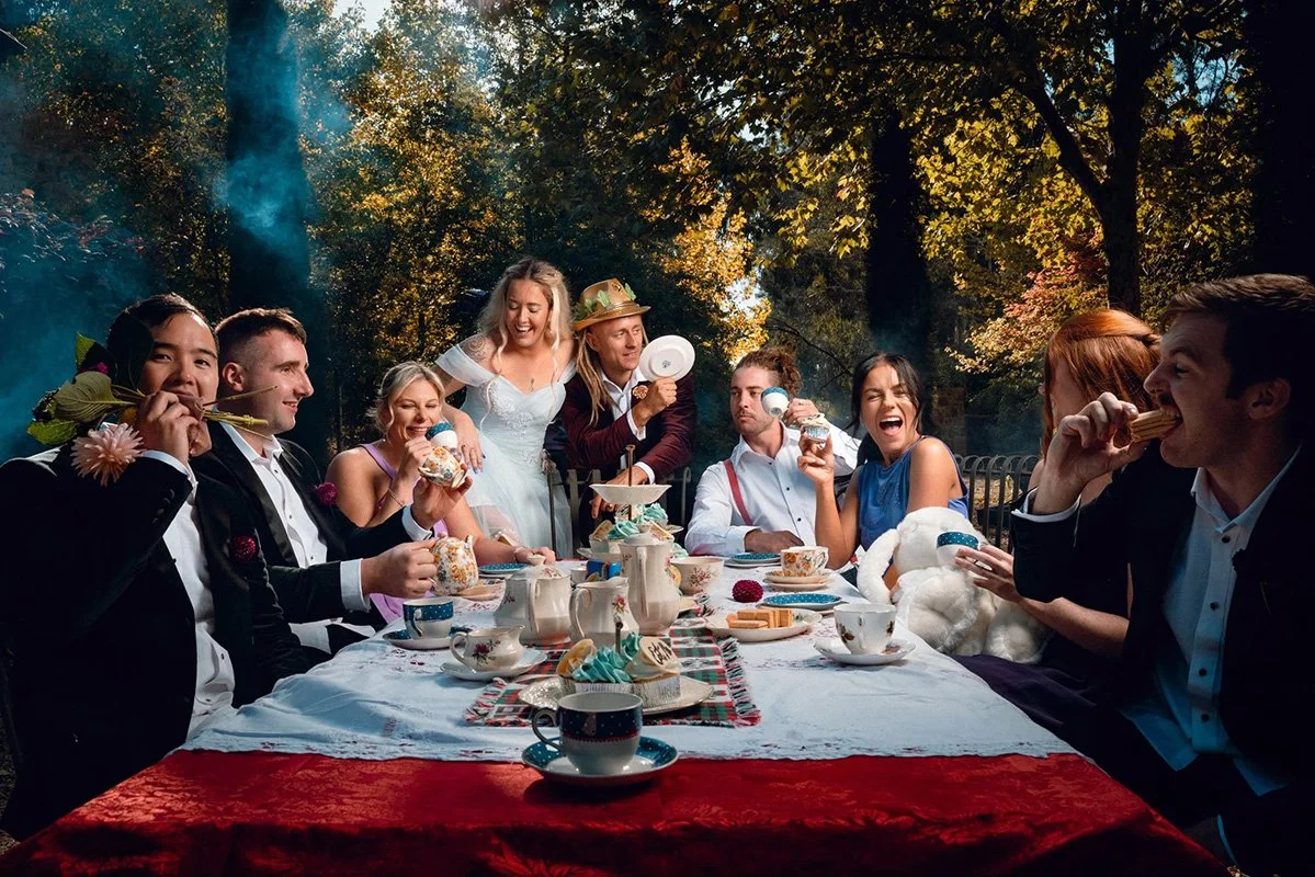 People at a festive outdoor tea party enjoying desserts, with a richly decorated table set in a wooded area during daytime.