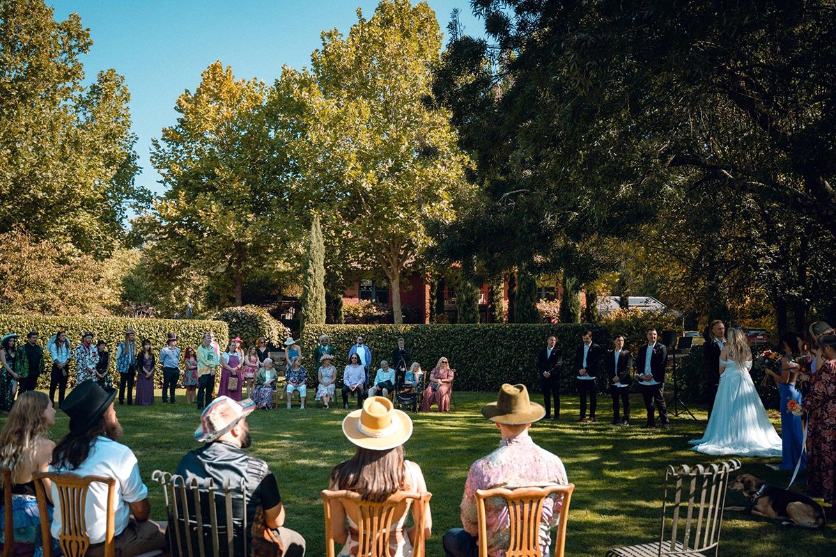 Outdoor wedding ceremony in a garden with brides and grooms, guests seated on chairs, and people standing under trees on a sunny day.