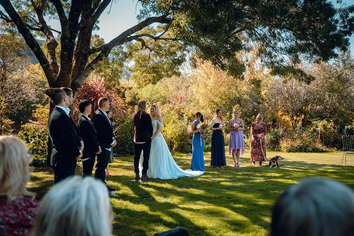 A wedding ceremony taking place outdoors in a garden with green grass and trees, featuring a bride in a white gown, a groom, and guests dressed in formal attire.