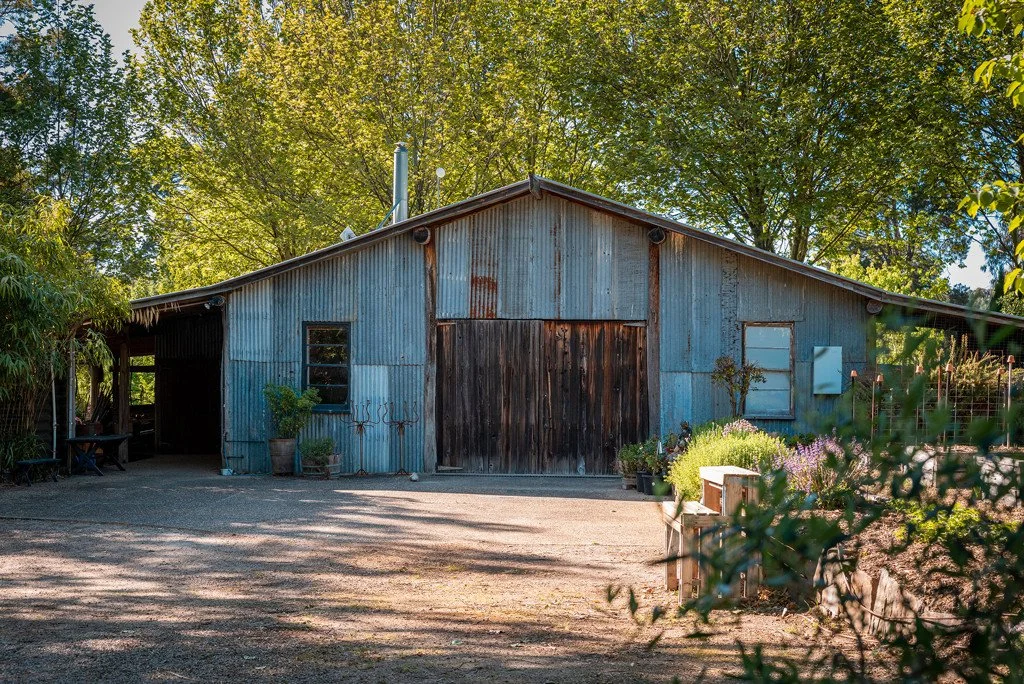 A weathered blue metal barn with wooden sliding doors, surrounded by trees and plants, with a gravel driveway in the foreground.