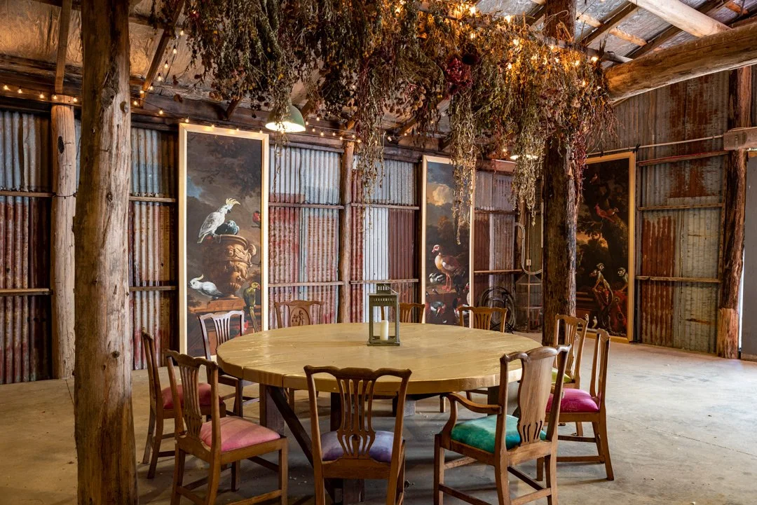 Rustic interior with wooden walls and round dining table surrounded by wooden chairs with colourful cushions, hanging dried flowers, and decorative paintings of animals.