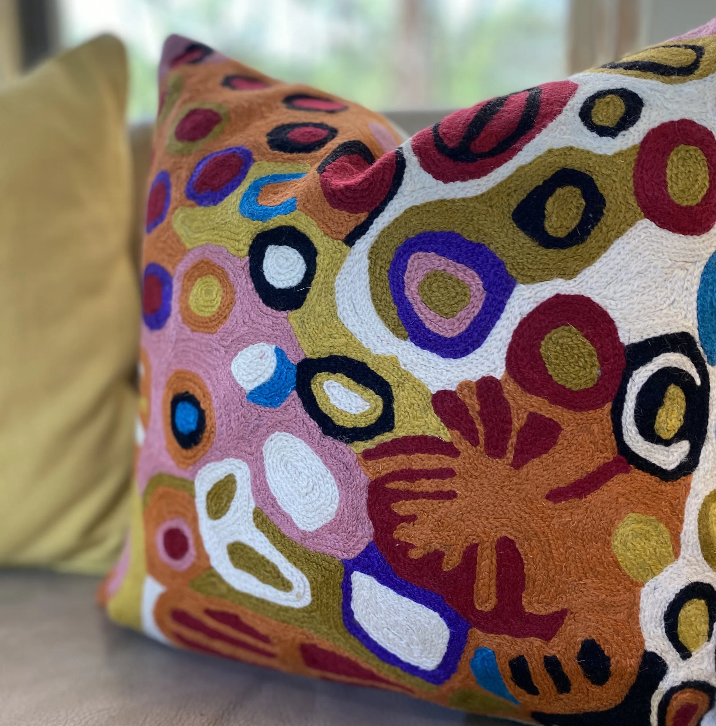 Close-up of a decorative pillow with colorful, abstract circular patterns, including red, purple, white, black, pink, blue, and yellow, on an indoor sofa with a window in the background.