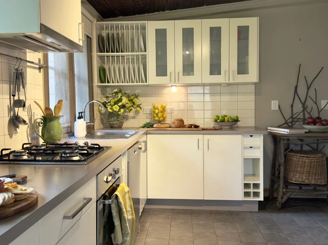 A kitchen with white cabinets, a countertop with a sink, a gas stove, and various decorative items including a vase with flowers, a bowl of lemons, and a plate of limes. There are also utensils hanging on the wall, a window, and a small table with a 