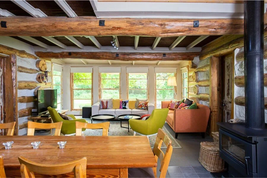 Living room with log cabin walls, wooden beams, large windows, and cozy furniture including a sofa, armchairs, and a wood stove.