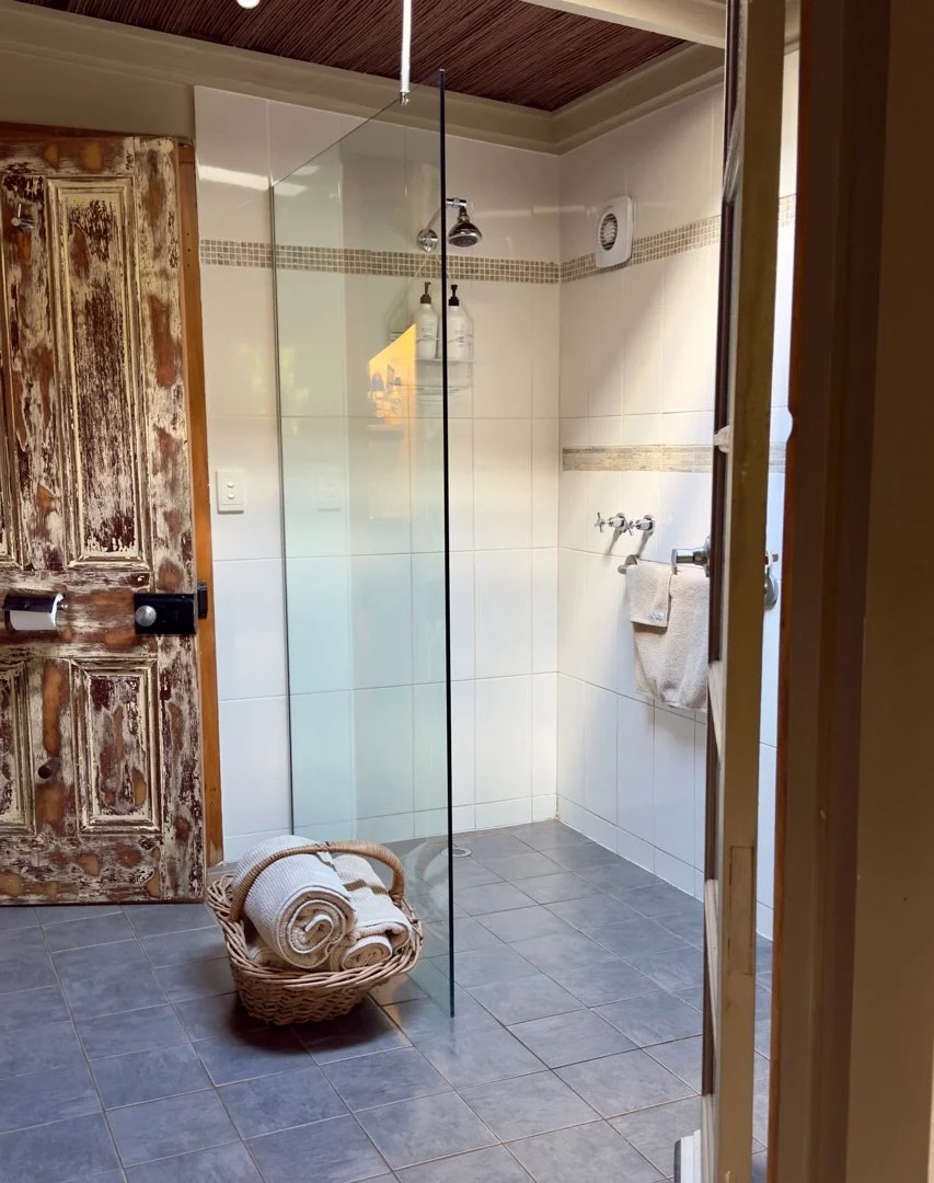 A bathroom with a walk-in shower area separated by a glass partition, featuring a rain showerhead, two bottles of shower products, a towel rack with a towel, a basket with rolled towels, and a rustic wooden door with peeling paint.