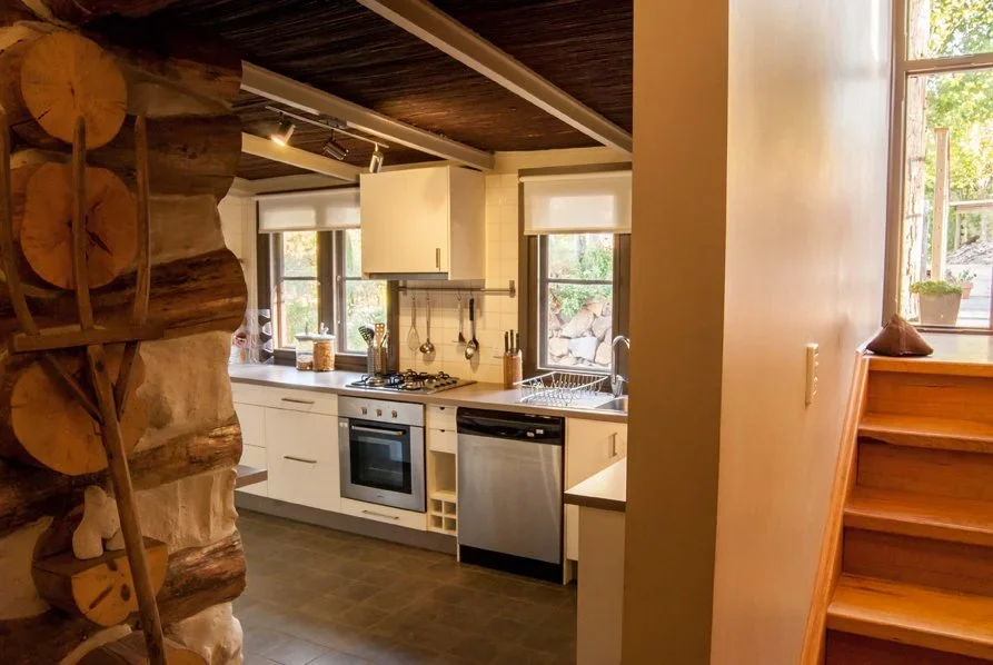 Cozy kitchen with white cabinets, a stainless steel dishwasher, a small oven, and a stove, with natural light from windows, and wooden stairs nearby.