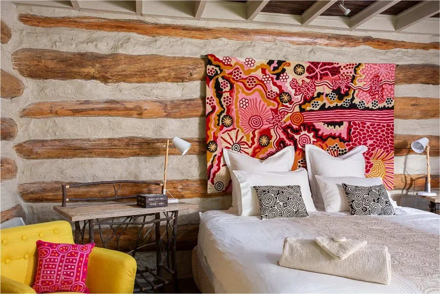 A rustic bedroom with exposed wooden beams and stone walls, featuring a colorful abstract tapestry above the bed, white bedding with decorative black and white pillows, a yellow armchair with a pink pillow, and two bedside lamps.