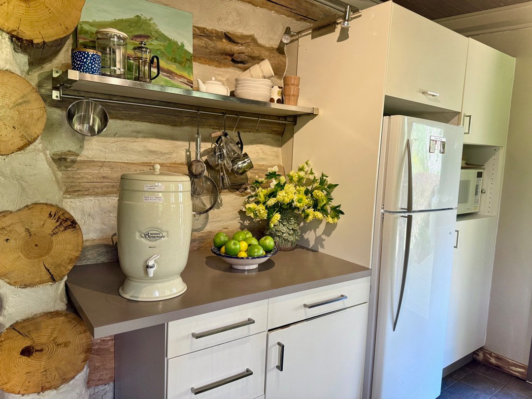 A kitchen counter with a vintage water dispenser, a bowl of green apples, and a bouquet of white flowers, with a white refrigerator and microwave in the background.