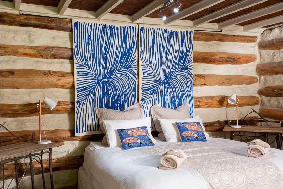 A cozy bedroom with a log cabin style wall, featuring a large blue abstract flower headboard, and a bed with white, beige, and patterned pillows, and rolled towels on the bed. There are two wooden nightstands with white lamps on each side.