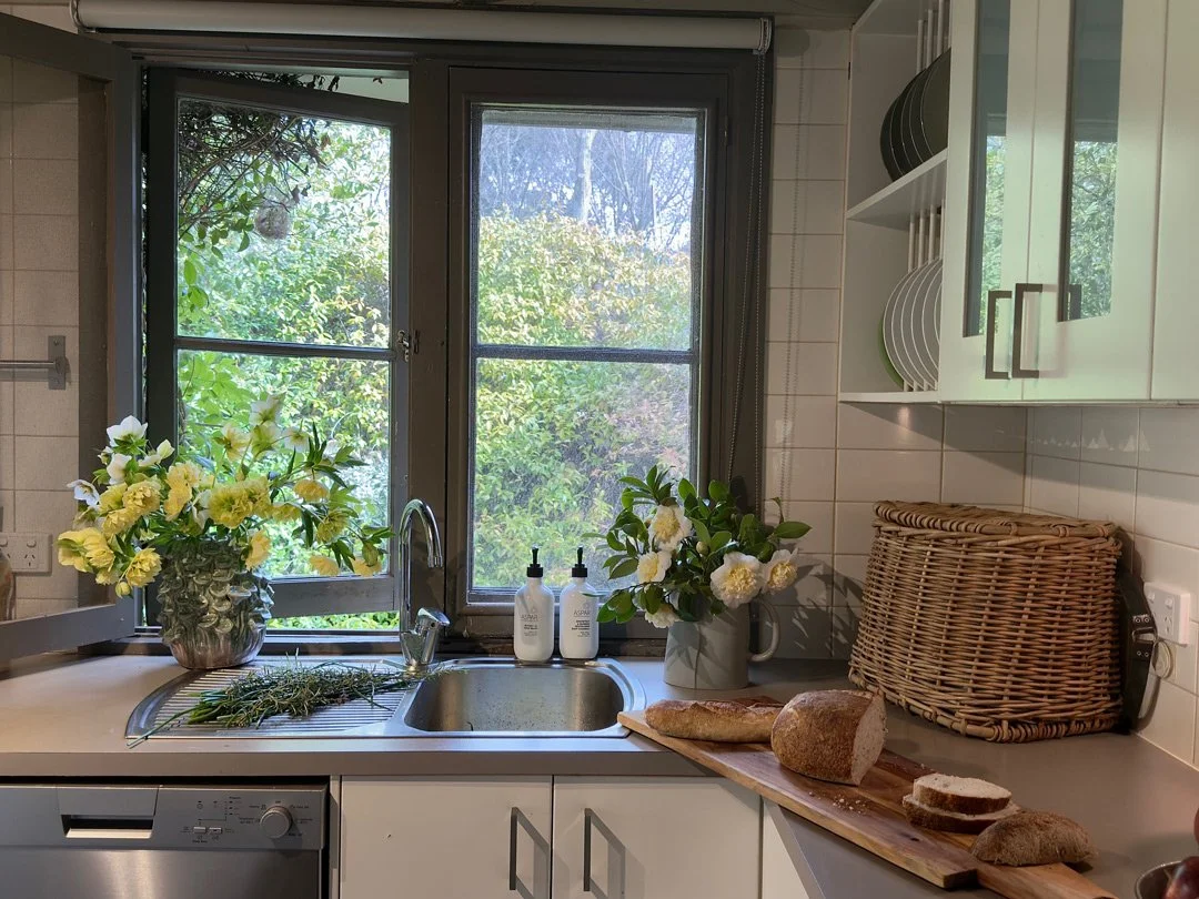 A kitchen with a window above the sink, showing greenery outside. The sink has a faucet with soap bottles and flowers on the windowsill. To the right, a cutting board with bread and loaf on it. There are white cabinets, a woven bread box, and dishware stored on open shelves.